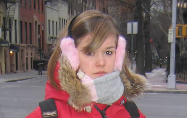 A girl in a red coat with pink fuzzy earmuffs and a gray and white scarf standing on a snowy street in New York