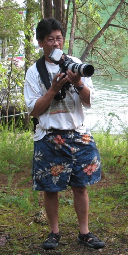A man in flowered shorts and a white t-shirt holding a camera with a large lens