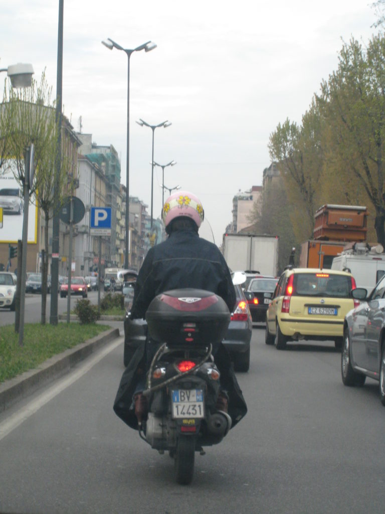 traffic in Milan, foreground a scooter, rider wearing a helmet decorated with daisies