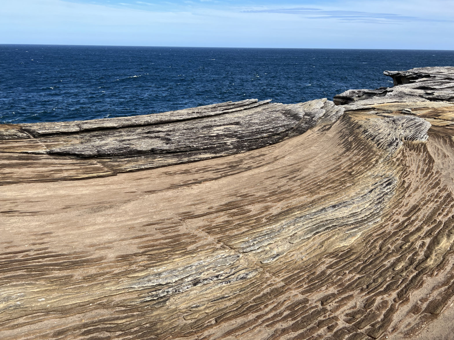 sandstone sculpted by wind and rain into wave forms, in shades of rust, beige, and ochre, with deep blue ocean and a light blue sky beyond