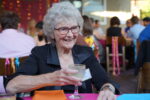 An older lady with curly gray hair and glasses grins as she holds a glass with a sugared rim, sitting at a colorful table