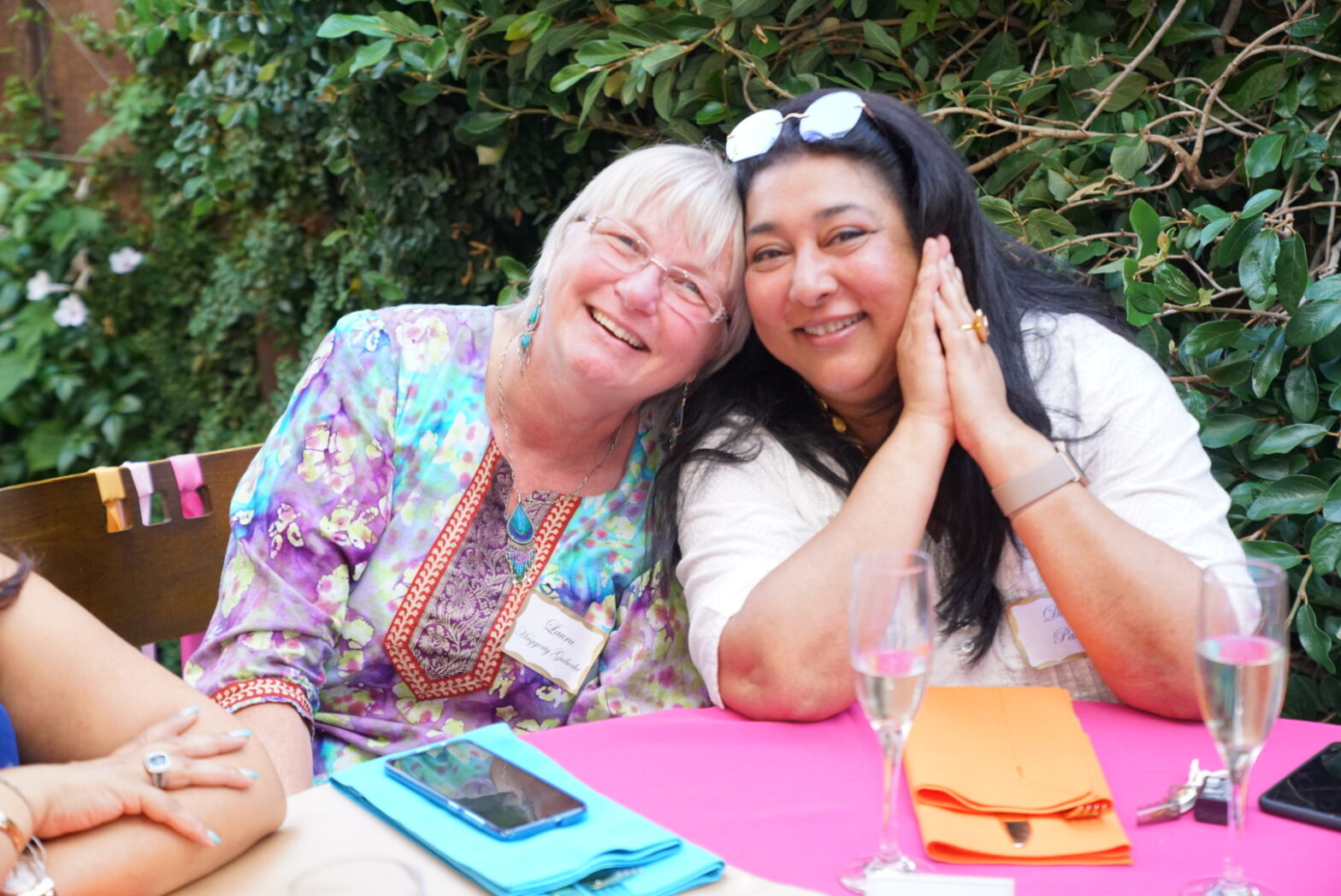 A Canadian woman with short white hair and an Indian woman with long black hair sitting at a table, leaning their heads together and smiling like the old friends they are