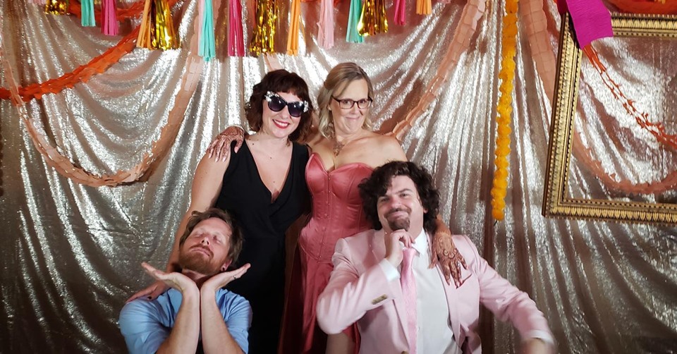 a man with his hands splayed out under his chin and eyes closed with a face of bliss, a woman wearing fancy sunglasses, a woman in a pink corseted wedding dress, a man in a pink jacket posing with his hand under his chin