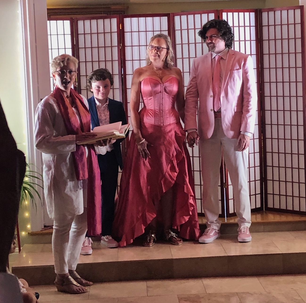 Woman on the left (Sue, the officiant) holds a book and looks into the crowd, making a statement or asking a question. Small boy looks down shyly. Bride and groom look at the officiant.