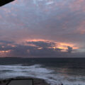 a cloudy, colorful sunrise over the smaller Cronulla ocean pool, which appears as a rectangle of black rock filled with still water, pointing towards the horizon. Beyond is the white water of breaking waves, and dark blue water in the pre-dawn light