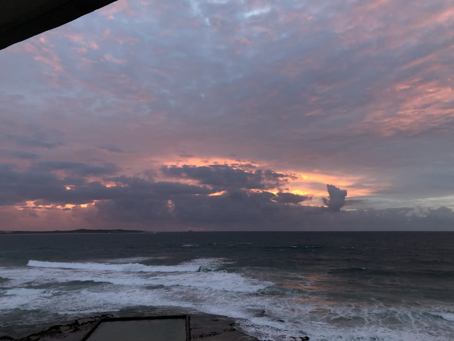 a cloudy, colorful sunrise over the smaller Cronulla ocean pool, which appears as a rectangle of black rock filled with still water, pointing towards the horizon. Beyond is the white water of breaking waves, and dark blue water in the pre-dawn light