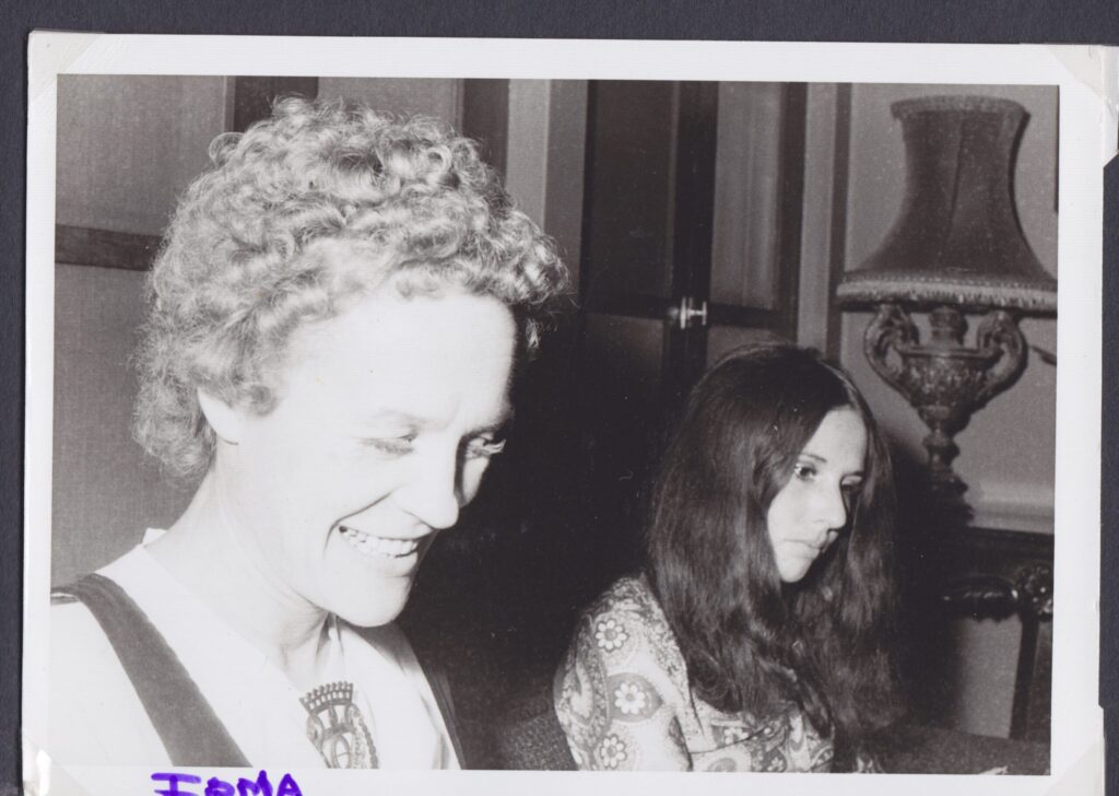 B&W - in the foreground a woman looks down, smiling. She has curly light hair. Beyond her is another woman with dark mid-length hair loose over her shoulders, looking abstracted