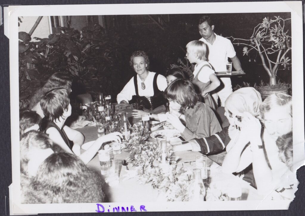 B&W - a long table of guests at a Christmas party. Irma with short curly hair wearing a Finnish traditional costume, seated at the end of the table, looks to the camera. Her daughter Hannah stands next to her, hands on hips, apparently berating. Behind Hanna is a Thai man with a glass of something on a tray. Other guests eat and look