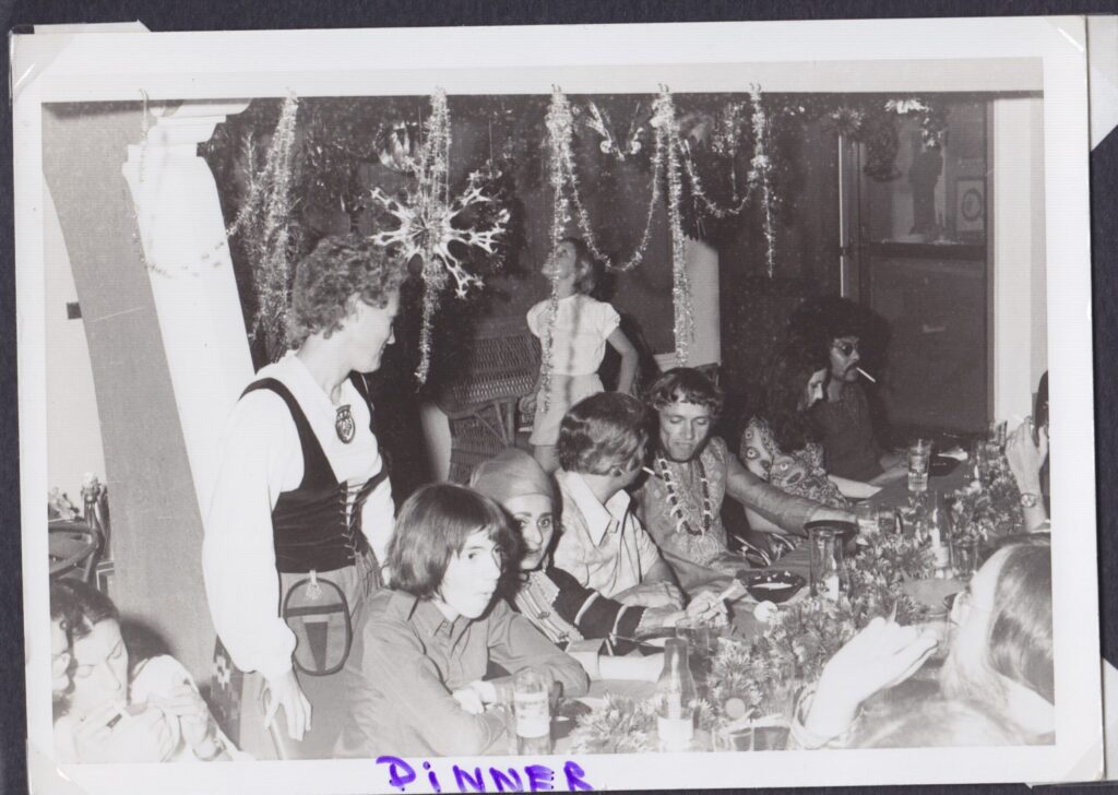 B&W Dinner table with various Christmast guests, tinsel streamers and stars hang form the ceiling. A black man at the end of the table with an afro wears sunglasses and has a cigarette hanging out of his mouth. Another (white) man has a necklace of dark beads and white animal fangs or claws. Irma stands overlooking the table