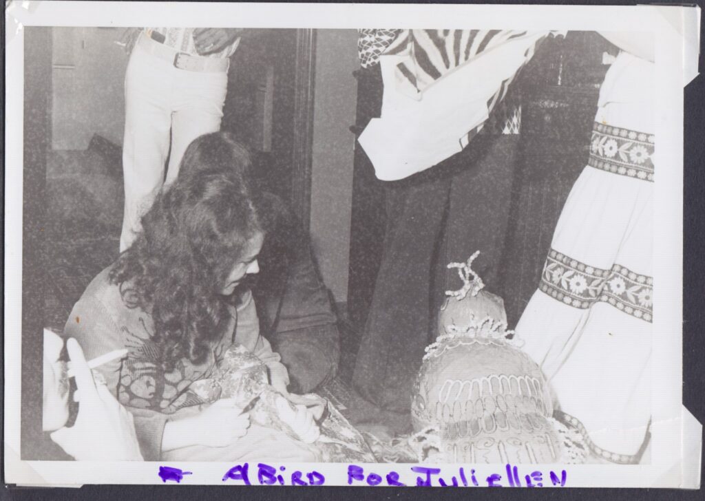 B&W - a woman sitting on the floor looks at a papier mache bird painted and decorated with beads that has been gifted to her for Christmas. In the foreground a hand holding a cigarette, other people seen from the waist down in the background wearing hippie fashions - one pair of white trousers, one pair of (probably jeans), one "peasant" skirt