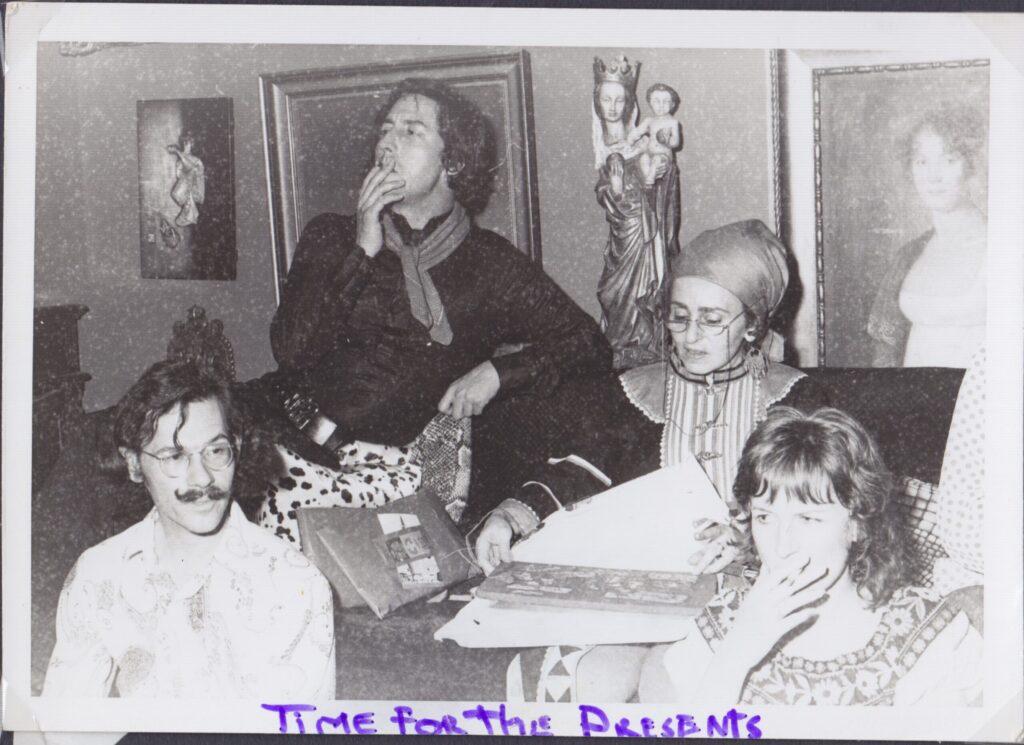 B&W - two men and two women sit on or in front of a sofa. A woman with glasses and a headscarf on is opening a gift. The man in a dark shirt with black and white animal print trousers gazes into the distance, smoking a cigarette or a joint. A man with a mustache curled up at the ends looks the other way. A woman on the floor in a "peasant" dress or blouse looks pensive, cigarette held to her mouth