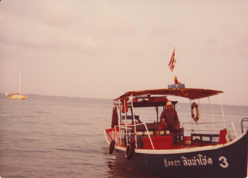 a Thai fishing boat with low sides, painted blue with white trim and an orange control area in the center covered by an awning. the owner stands at the helm, gazing at the camera