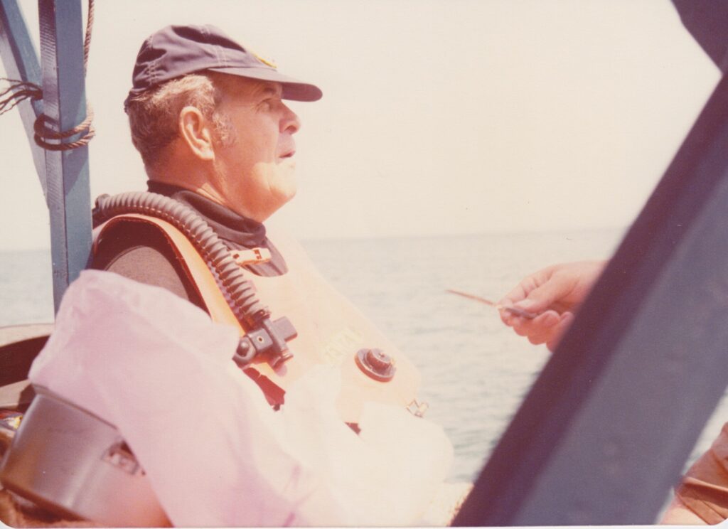 "Dusty" Rhodes, an older man with gray hair, wearing a slouchy, faded blue cap with a bill and an orange diving vest with an inflation tube going over his shoulder. someone off camera is gesturing towards him with a small knife (no, it was not a threat)