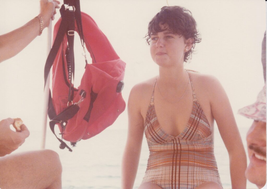 A girl with short curly brown hair in a bathing suit sits on the edge of the boat facing in. next to her an orange life vest hangs limply