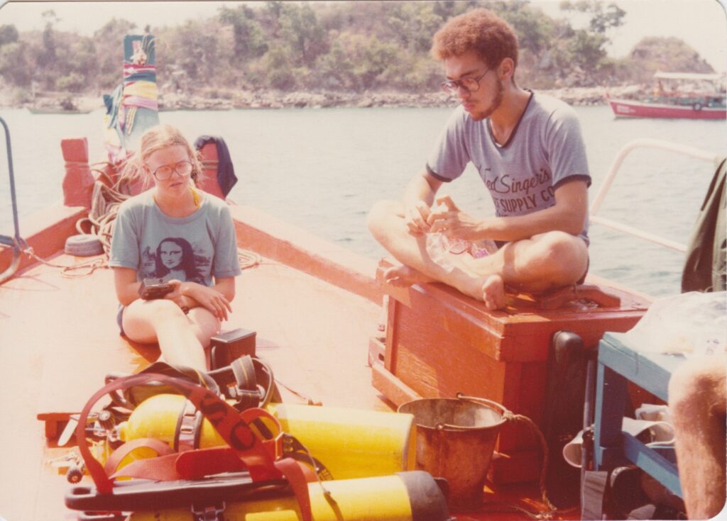 myself in a blue t-shirt seated on the foredeck of the boat with my lecgs stretched out in front of me, to the side a young man with curly dark hair sits cross-legged on a wooden chest. Between us are several yellow dive tanks and other gear
