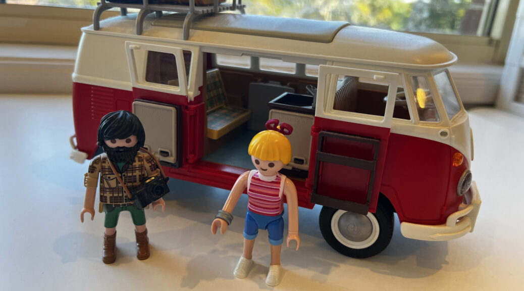toy model of a classic VW camper, with two figurines standing next to it. The male figure has shaggy black hair and a beard and mustache, the female is blonde