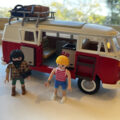 toy model of a classic VW camper, with two figurines standing next to it. The male figure has shaggy black hair and a beard and mustache, the female is blonde